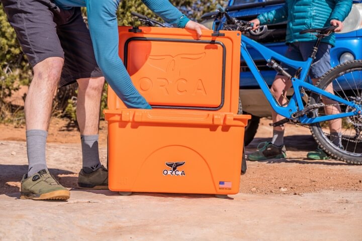 An orange cooler with a person (only their legs, arms and small part of torso are in the frame) bent over reaching into the cooler. In the background another person standing next to bike with hands on the seat and handlebar.
